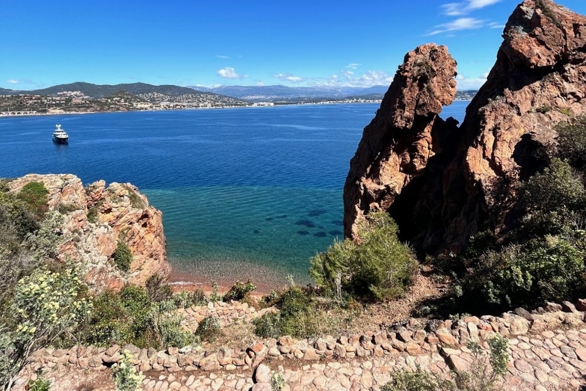A stone path between red-rock pinnacles at the Pointe de l'Aiguille, with a turquoise cove below and the bay of Cannes on the horizon