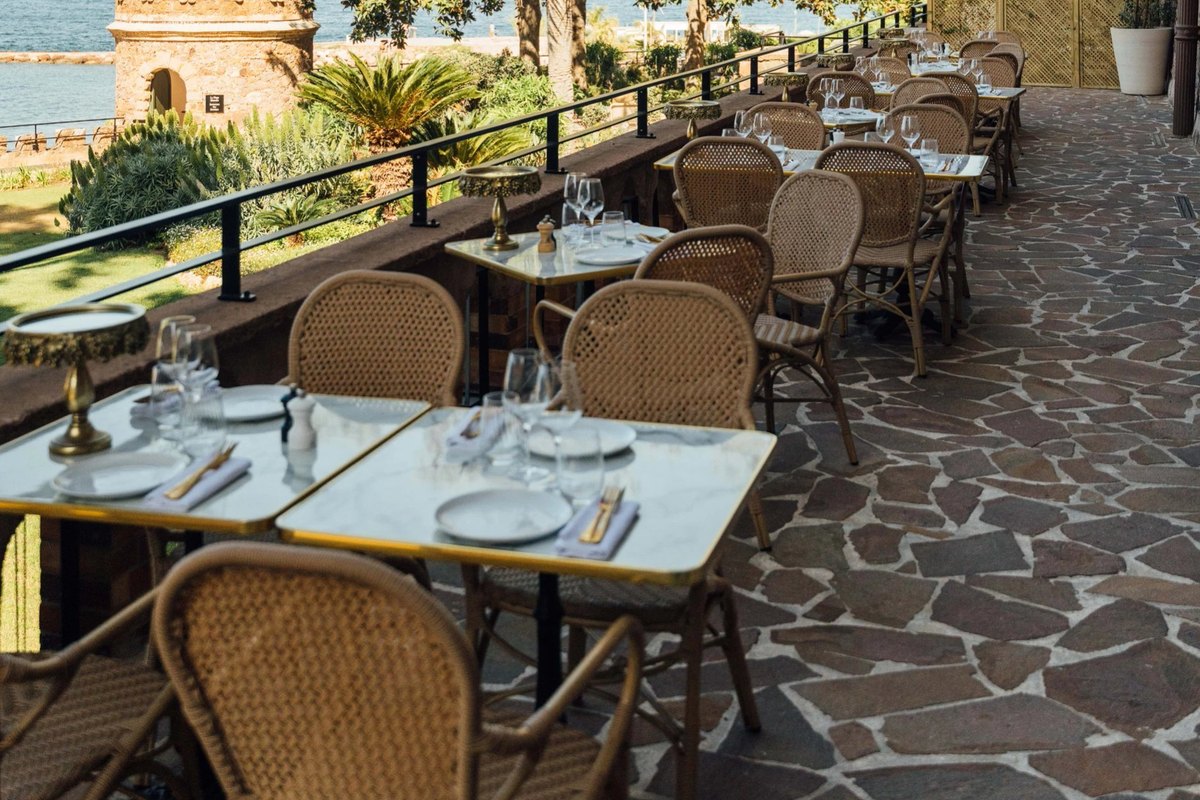 Rattan chairs and laid tables on the Château de Théoule terrace along the ramparts, with the sea beyond the railings