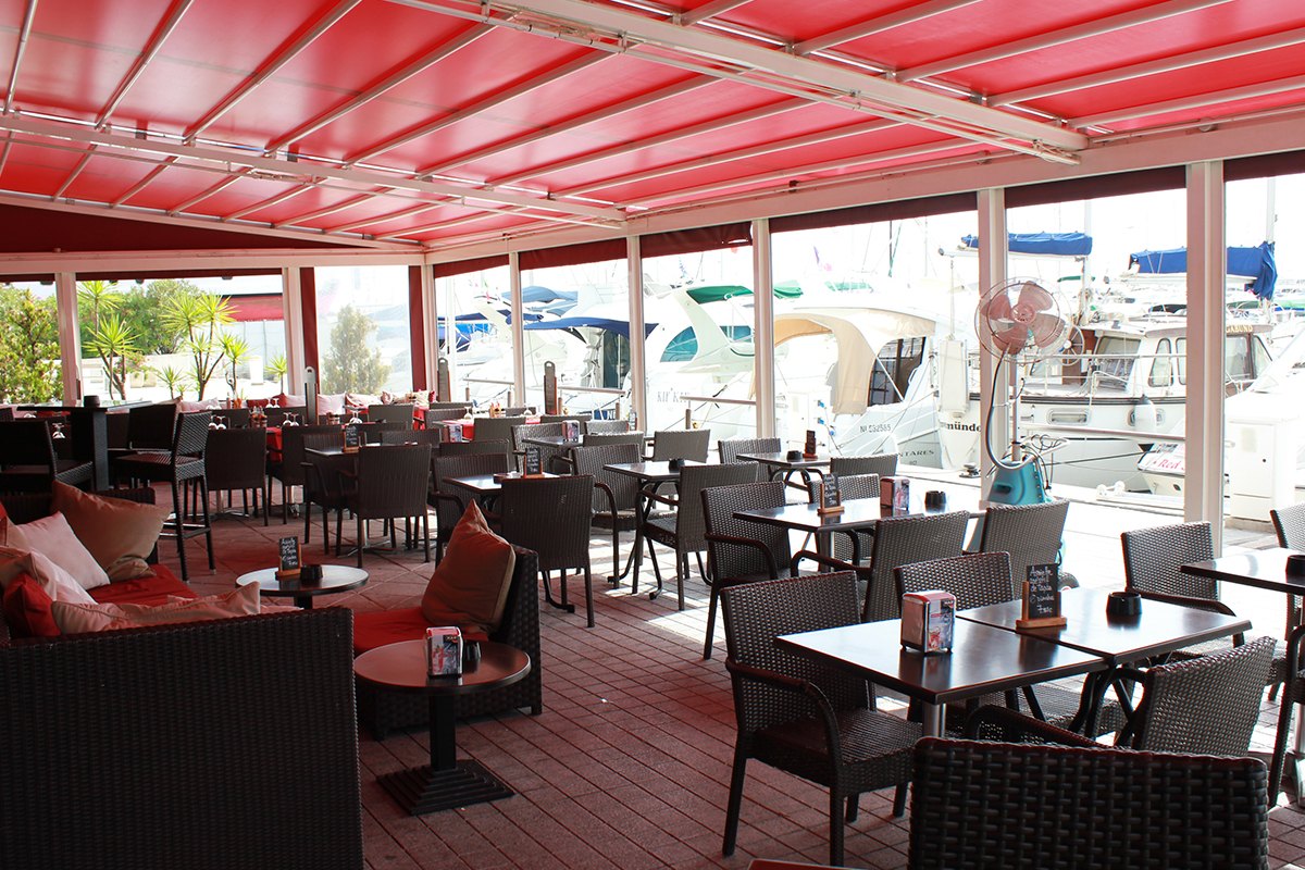 The covered terrace of Rio's Banana Café under red awnings, looking out over the yachts of Port Camille Rayon