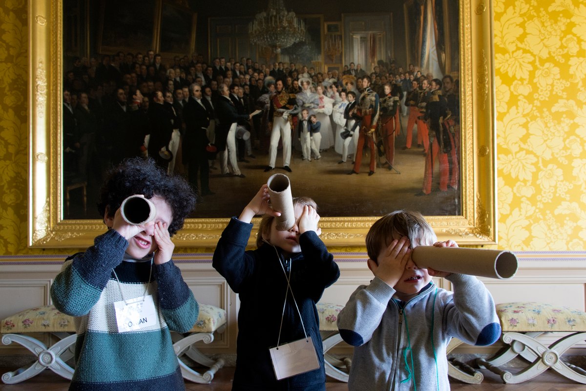 Three children peering through paper telescopes in front of a gilded painting during a family visit at the Palace of Versailles