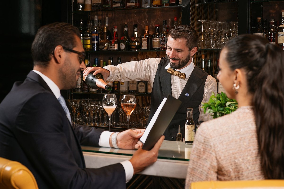 A bartender in a bow tie pours wine for two guests at the Peacock Alley Bar Galerie, Waldorf Astoria Versailles – Trianon Palace