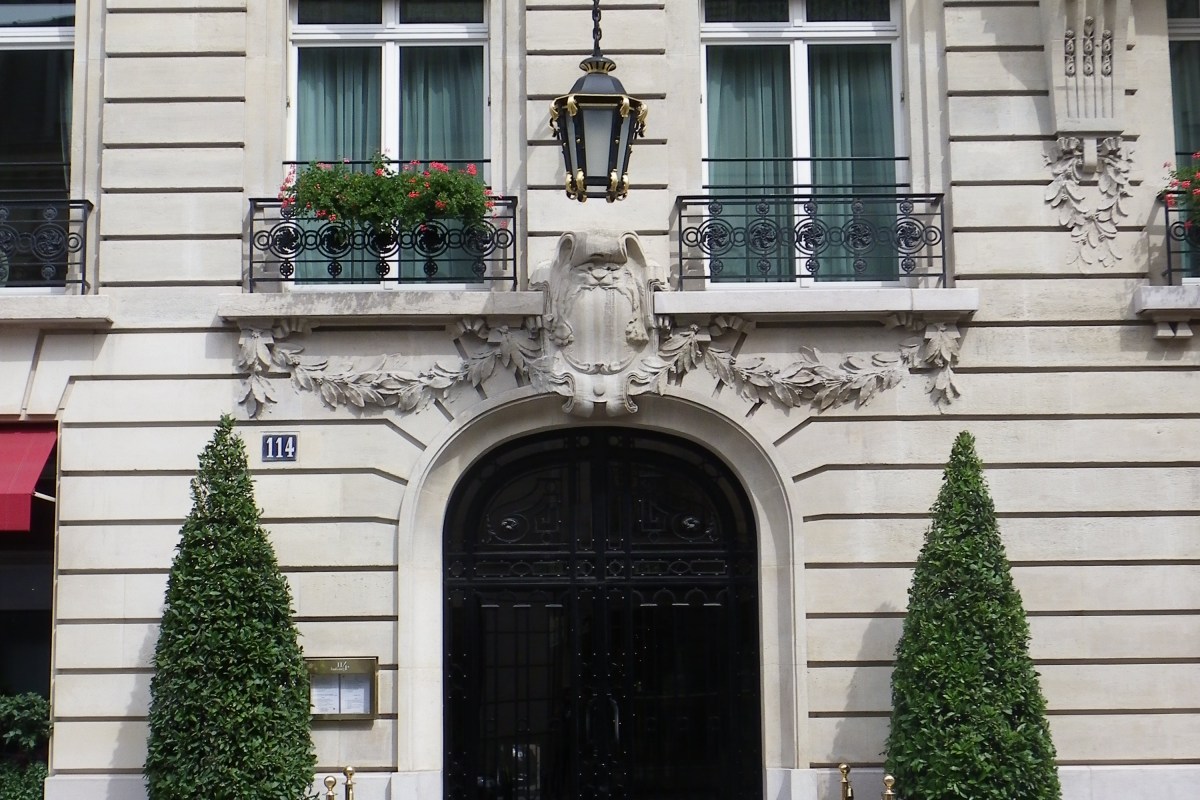 Ornate Haussmannian building facade with arched black entrance door, topiary trees, and '114' address plaque, rue du Faubourg-Saint-Honoré, Paris.