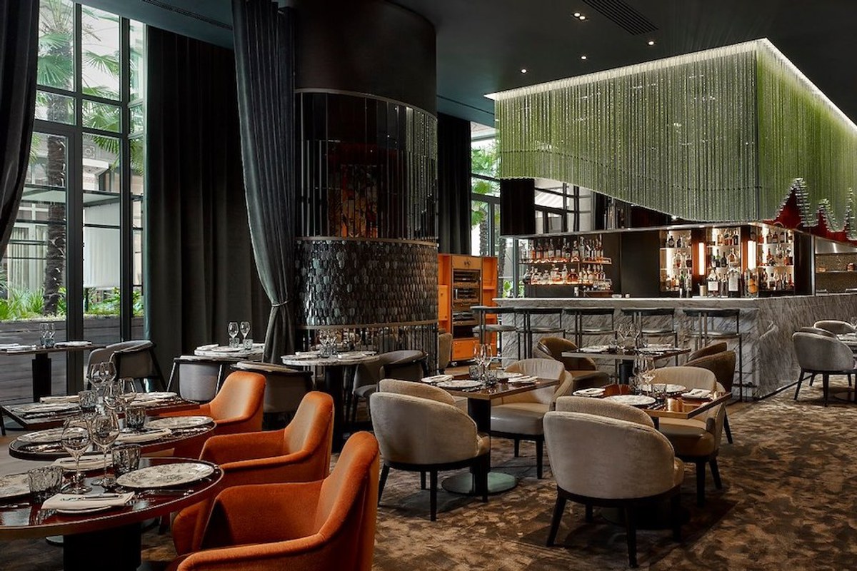 Restaurant dining room and bar with orange and grey chairs, green hanging light feature, floor-to-ceiling windows