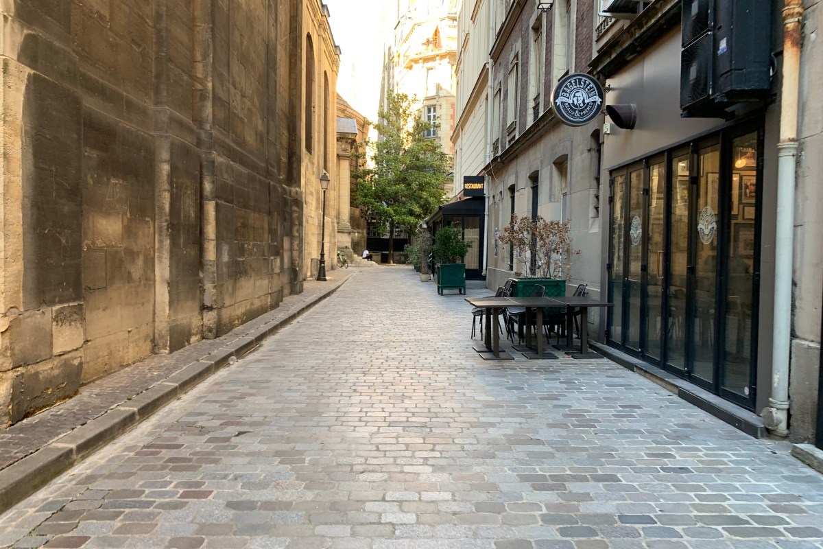 Cobblestone passage with a restaurant terrace table and chairs, awning and signage visible, church wall on left