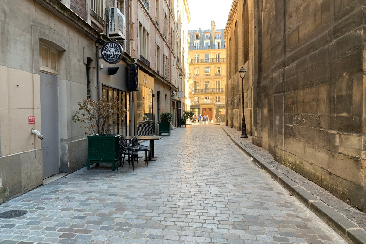 Cobblestone passage lined with shopfronts, a terrace table set outside, church wall on the right