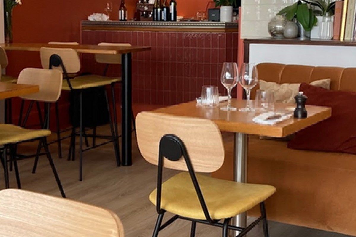 Restaurant dining room interior with wooden tables, yellow chairs, orange banquette and tiled bar area.