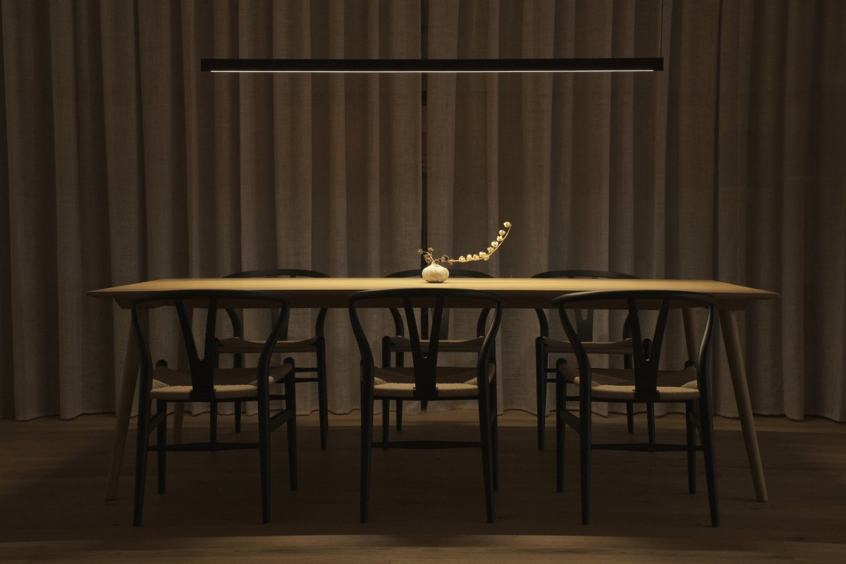 Long wooden dining table with black wishbone chairs under a linear pendant light against curtains