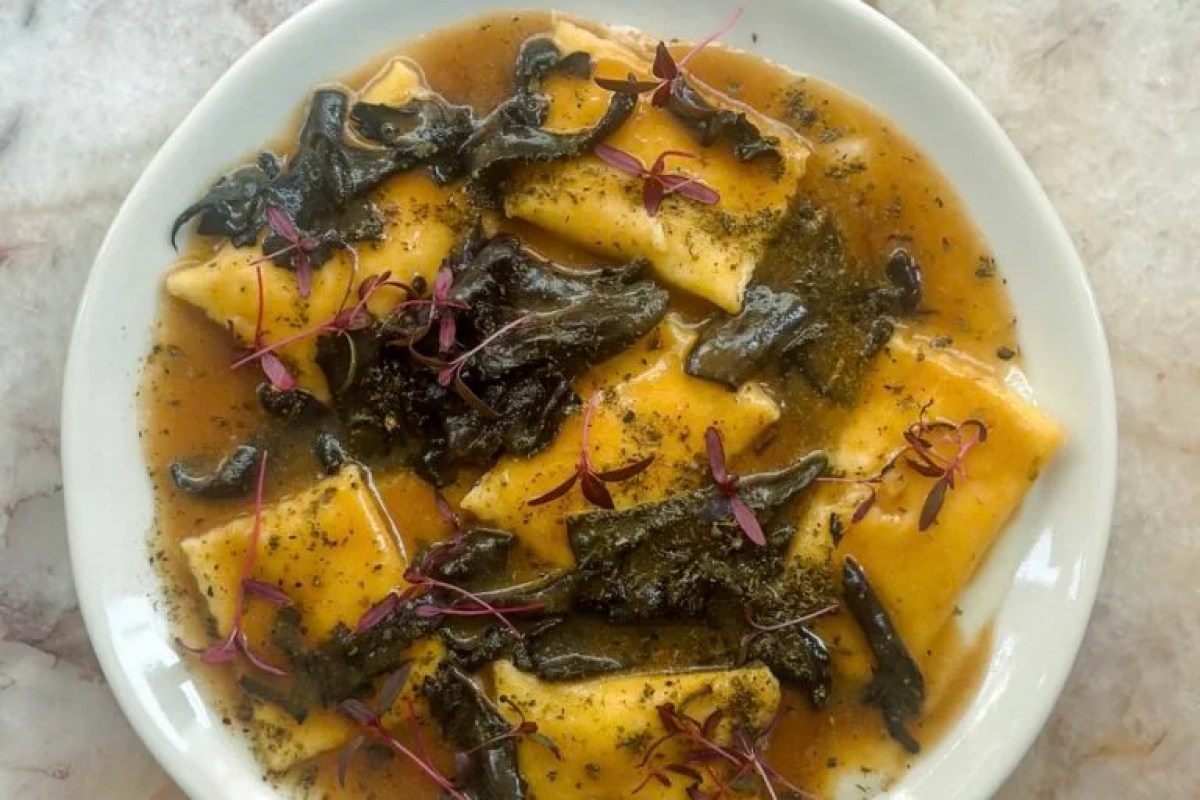 Overhead view of a plate with yellow ravioli in brown broth, dark mushrooms, and purple microgreens on a marble table.