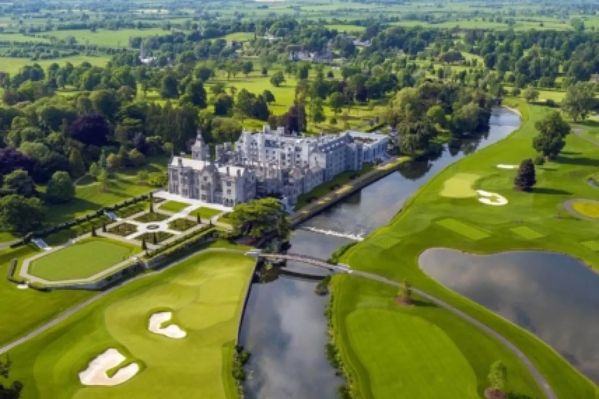 Aerial view of Adare Manor's grand stone façade beside a river, surrounded by golf course greens and lush parkland.