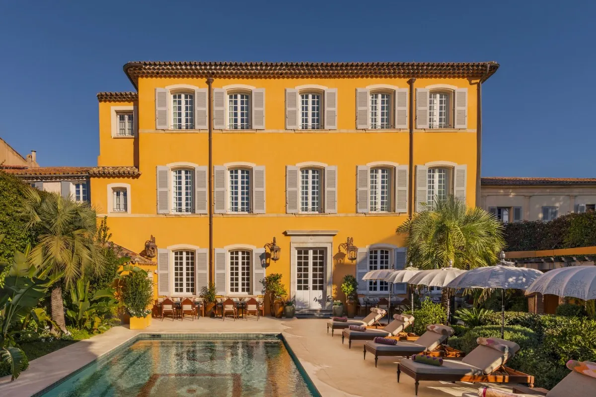 Ochre-yellow three-story hotel façade with shuttered windows, pool and lounge chairs in foreground
