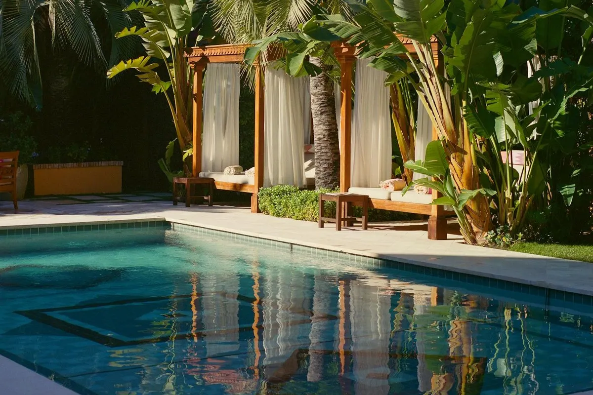 Hotel pool with wooden cabana daybeds shaded by curtains and palm trees