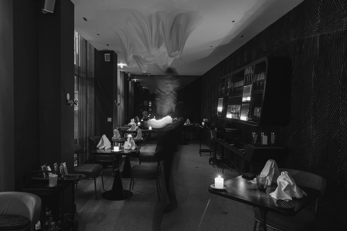 Dim dining room with candlelit tables, napkins, a bar shelf, and a blurred figure of staff walking