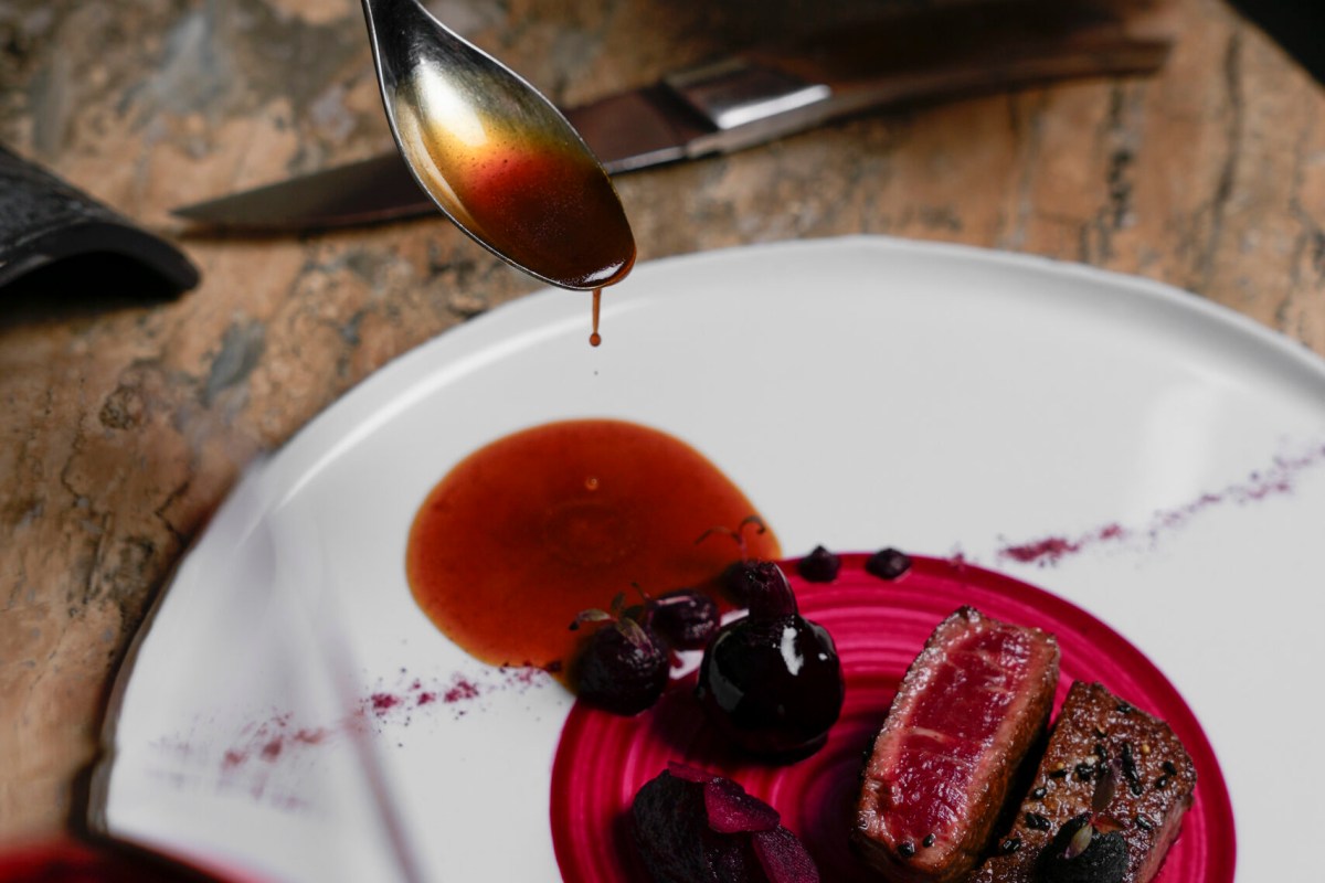 Plated dish with seared meat, dark cherries, and sauce being drizzled from a spoon on a white plate