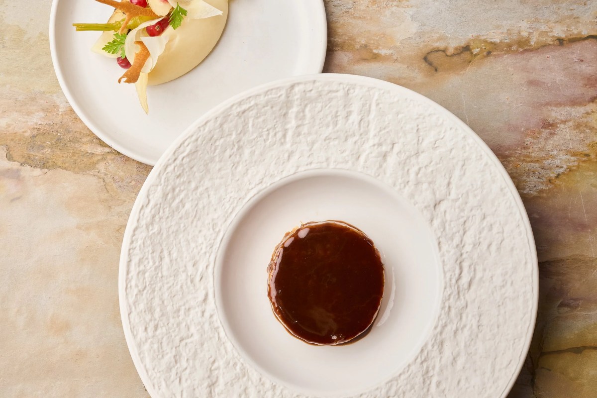 Two plated dishes on textured white plates atop marbled stone table, one with dark glaze, one with vegetables