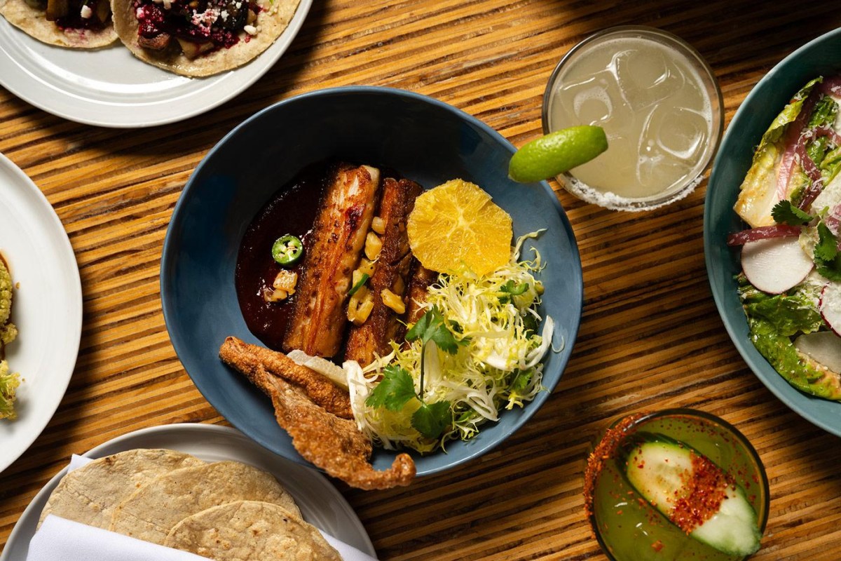 Overhead shot of pork rib dish with corn, citrus, frisée salad, and a cocktail on a wooden table