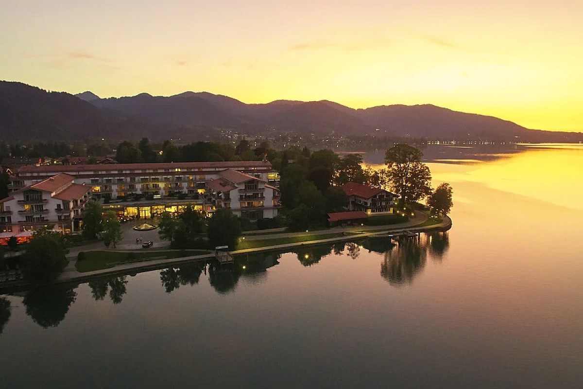 Aerial view of Seehotel Überfahrt at sunset, lakeside hotel building glowing with warm lights beside a golden lake