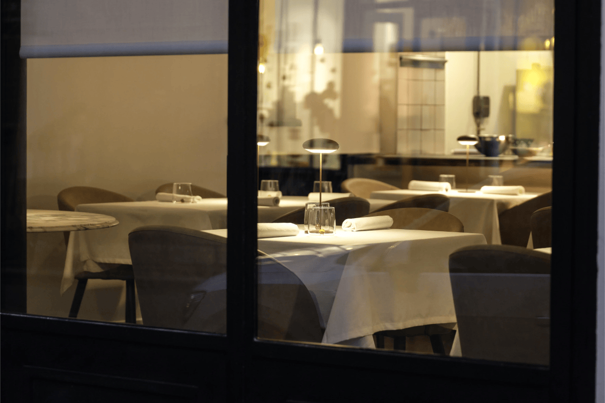 View through the restaurant's window at night showing the dining room with set tables, white tablecloths and soft lighting