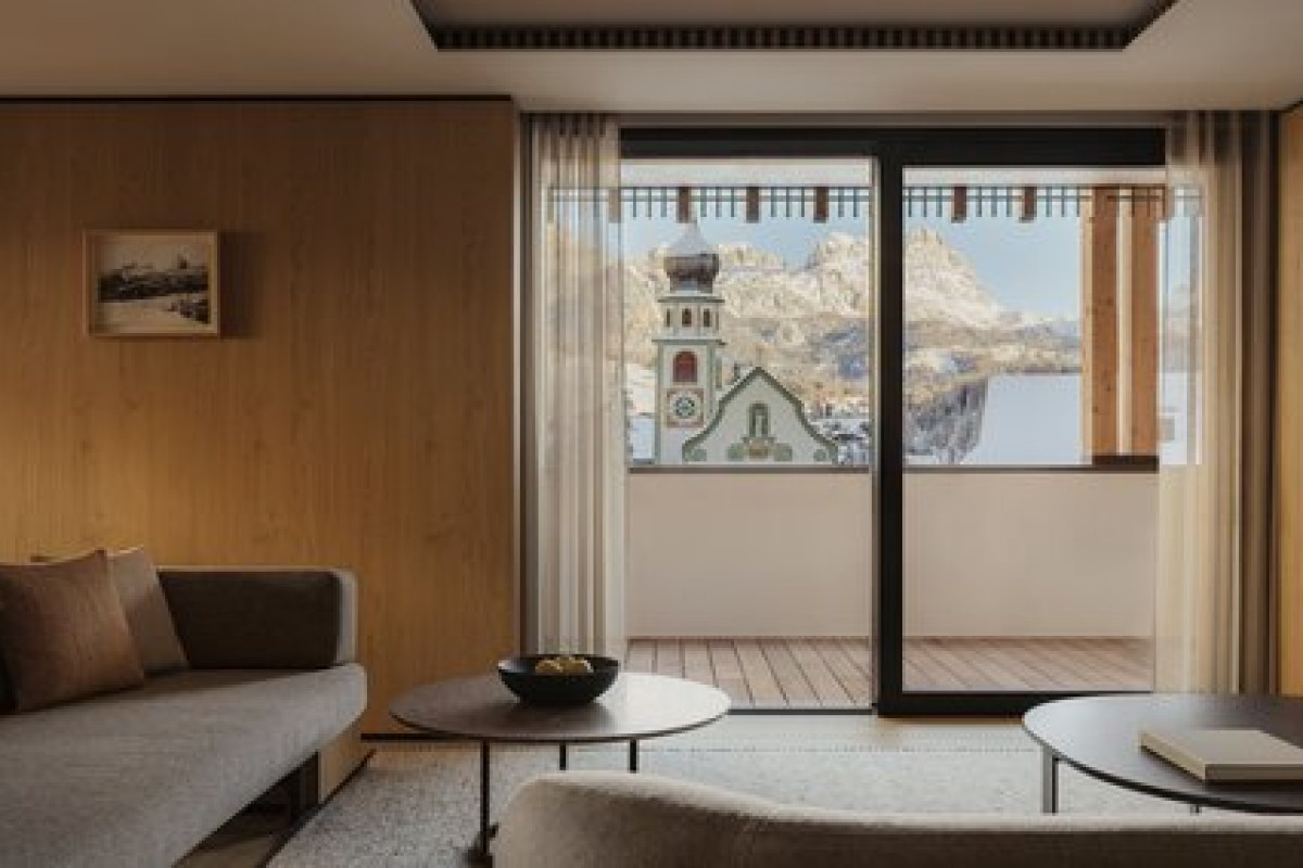 Guest room with wood paneling, sofa, and balcony view of a village church and Dolomites