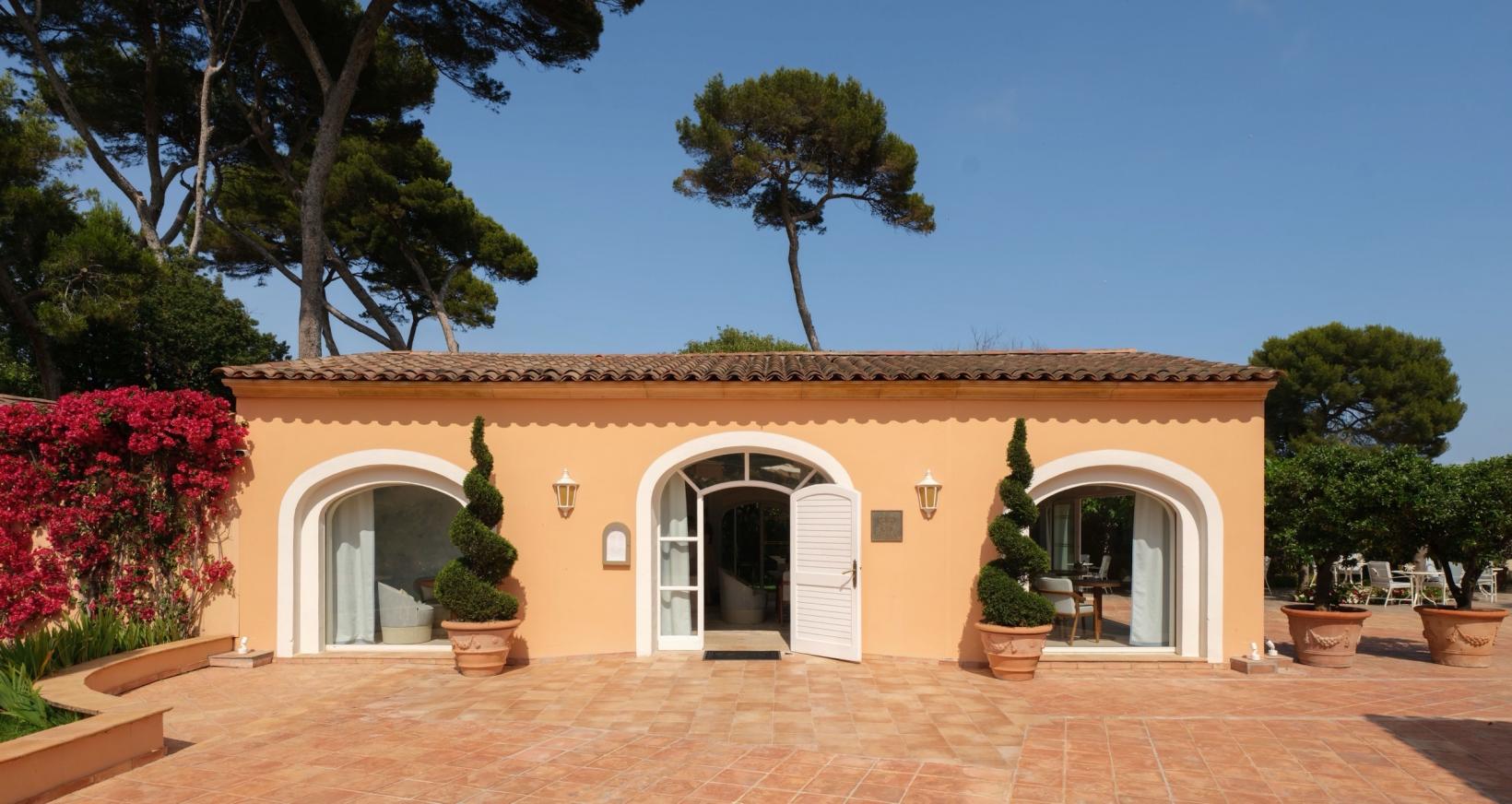 The garden pavilion of Amarines at Villa Miraé on Cap d'Antibes, an ochre building with arched doors, terracotta tiles, potted topiary and umbrella pines
