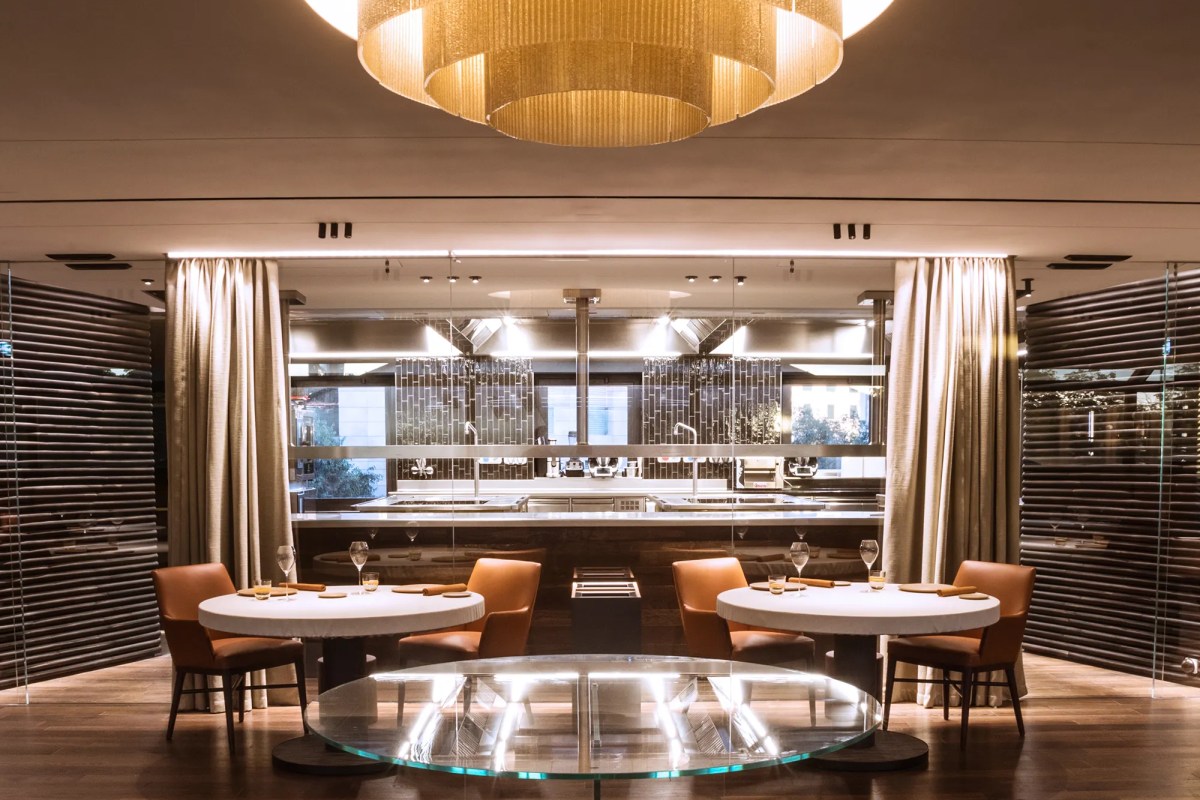 Elegant dining room with round tables, leather chairs, and a view into the open kitchen under a gold chandelier