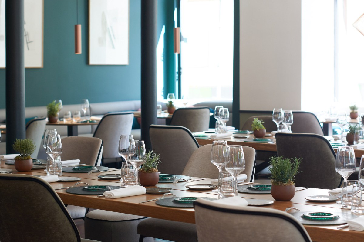 Restaurant dining room with wood tables set with glassware, small terracotta herb pots, and teal accent wall