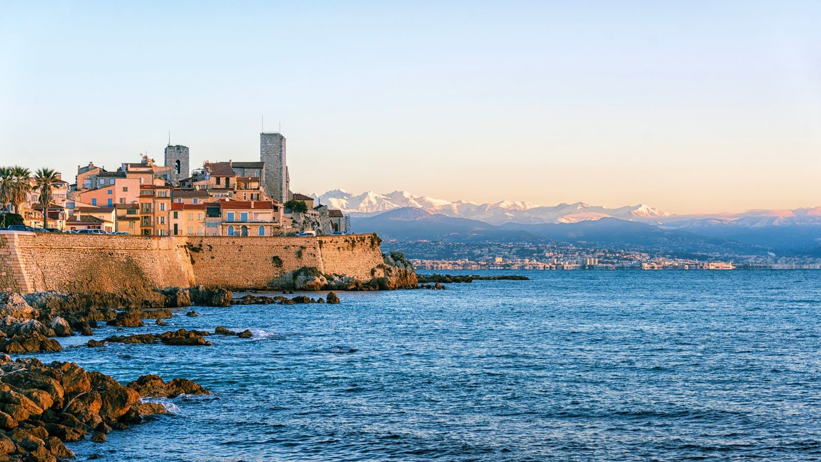 The old town of Antibes on its sea wall in golden evening light, with the Château Grimaldi tower and the snow-capped Alps across the bay
