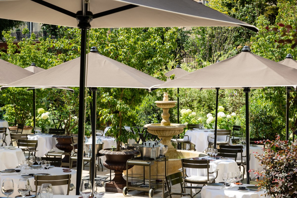 Elegant outdoor terrace with white-clothed tables, umbrellas, and a stone fountain surrounded by greenery.