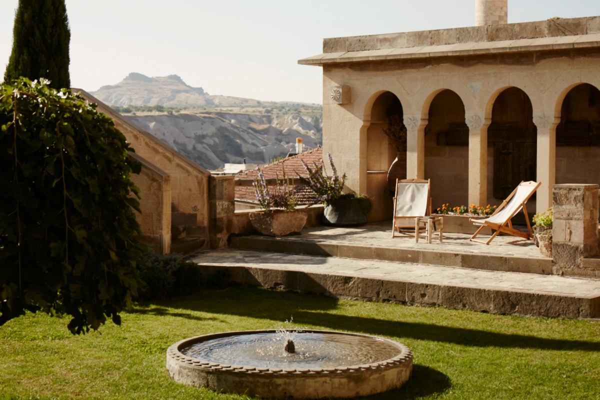 Stone hotel facade with arched terrace, garden chairs, and fountain on lawn, Cappadocia valley in background