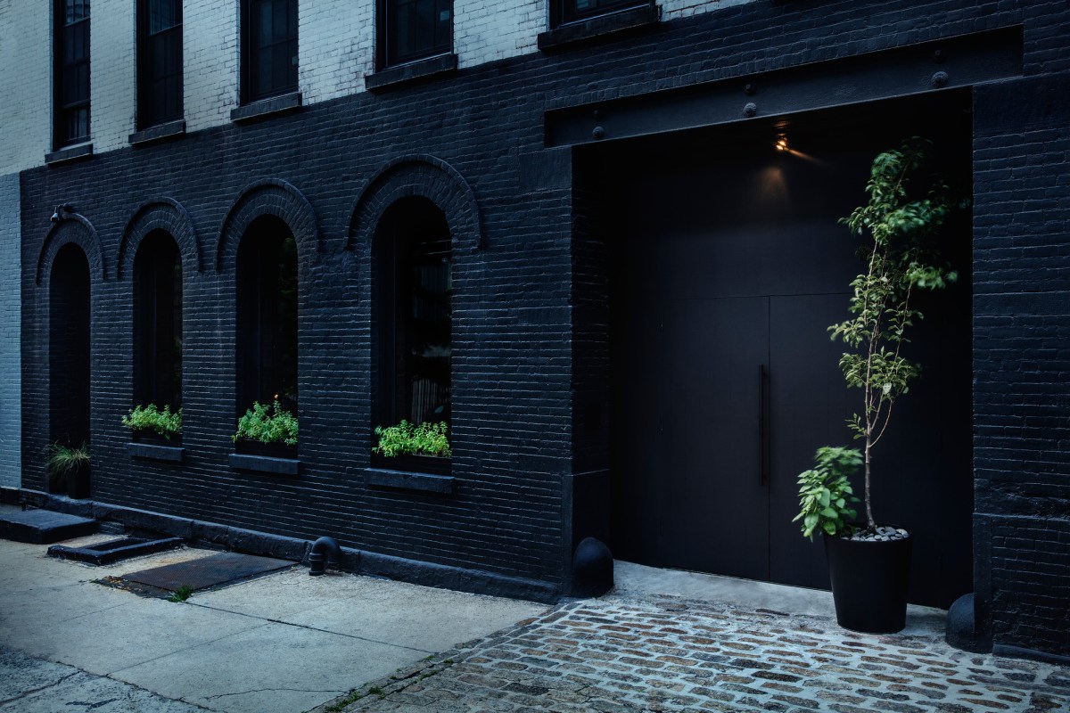 Exterior facade of Aska restaurant with black brick walls, arched windows with greenery, and dark entrance door flanked by a potted tree