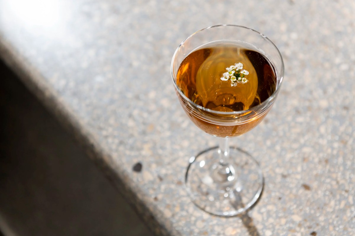 Amber cocktail garnished with a small white flower in a coupe glass on a terrazzo counter