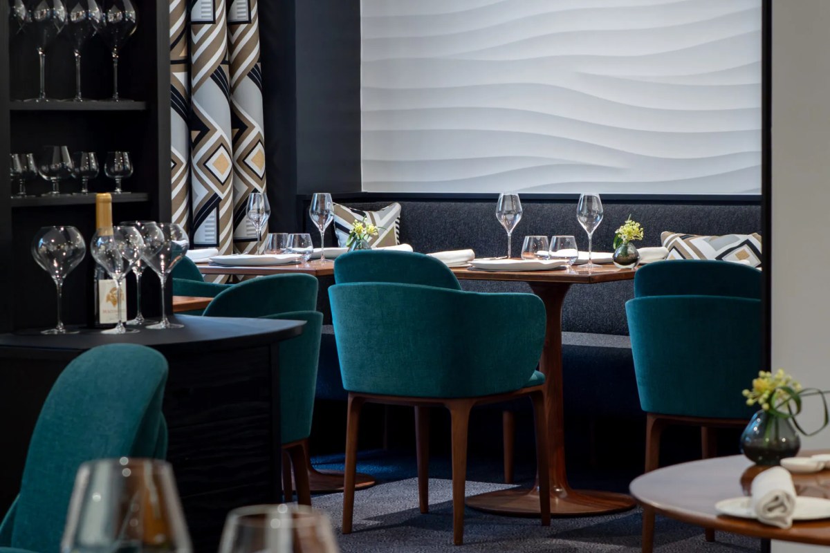Restaurant dining room with teal velvet chairs, wood tables, wine glasses and patterned curtains