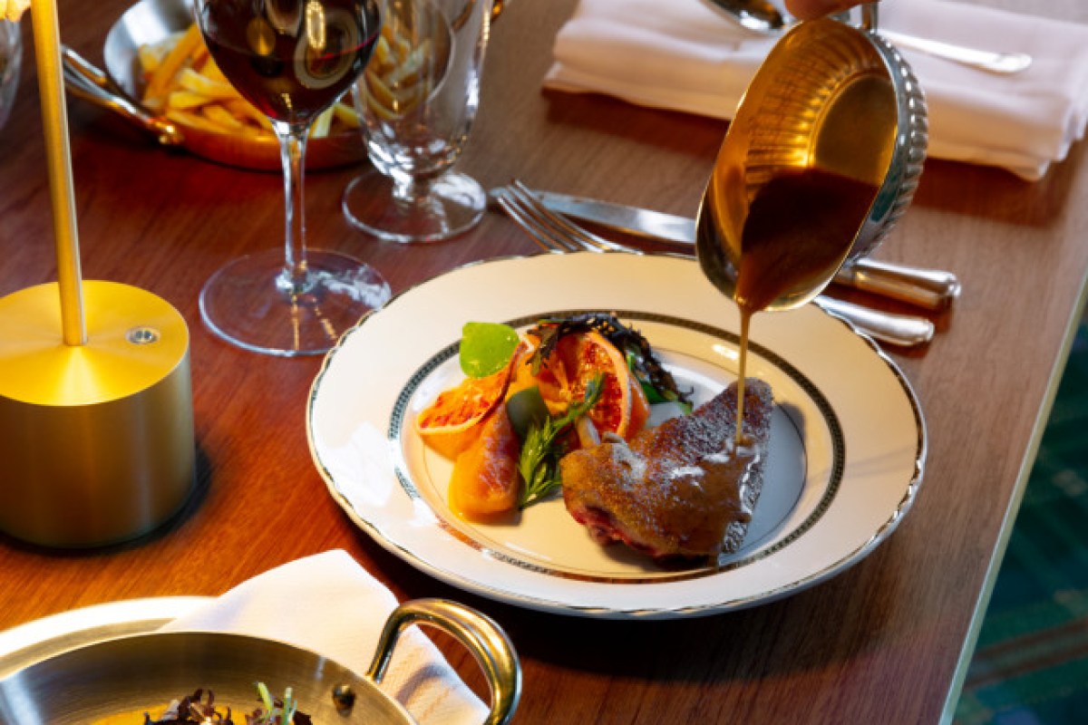 Plate of duck with citrus garnish, sauce being poured over it, wine and fries in background