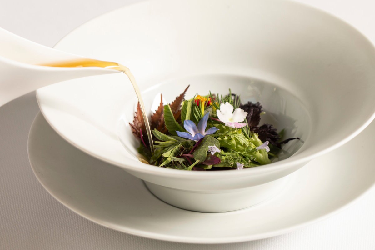 White bowl with a fresh herb and edible flower salad, olive oil being poured in, on a white table setting
