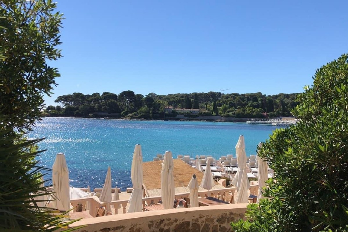 The terrace of the Hôtel Baie Dorée on Cap d'Antibes, with furled white parasols and a wooden deck above the turquoise Garoupe bay and its pine-covered shore