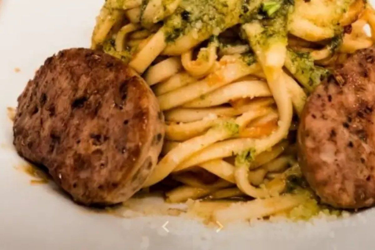 Pasta dish with pesto sauce and two slices of sausage on a white plate