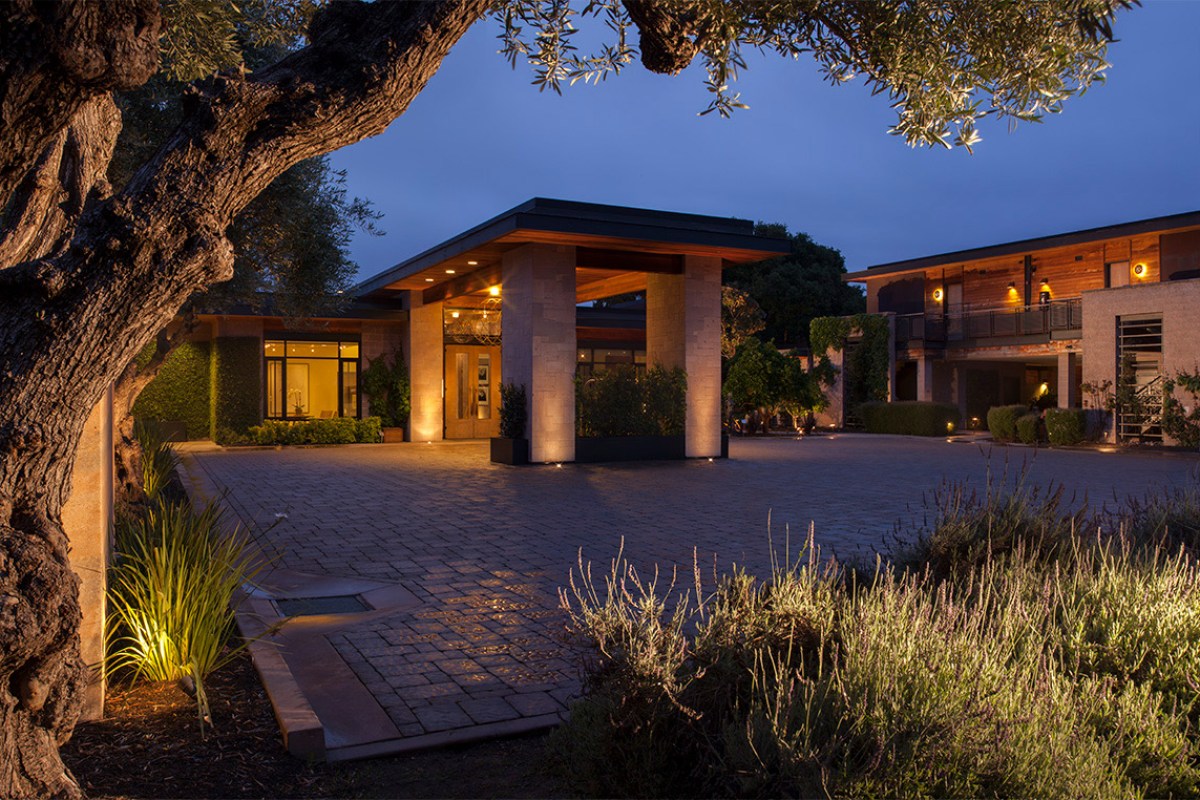 Hotel entrance at dusk with modern canopy roof, stone pillars, and warm lighting under an olive tree