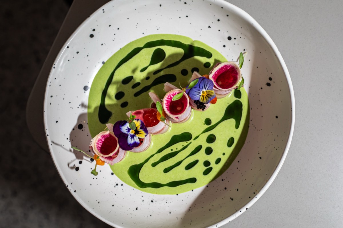 Overhead view of plated fish dish with pink radish cups, edible flowers, and green sauce swirls on a speckled white plate