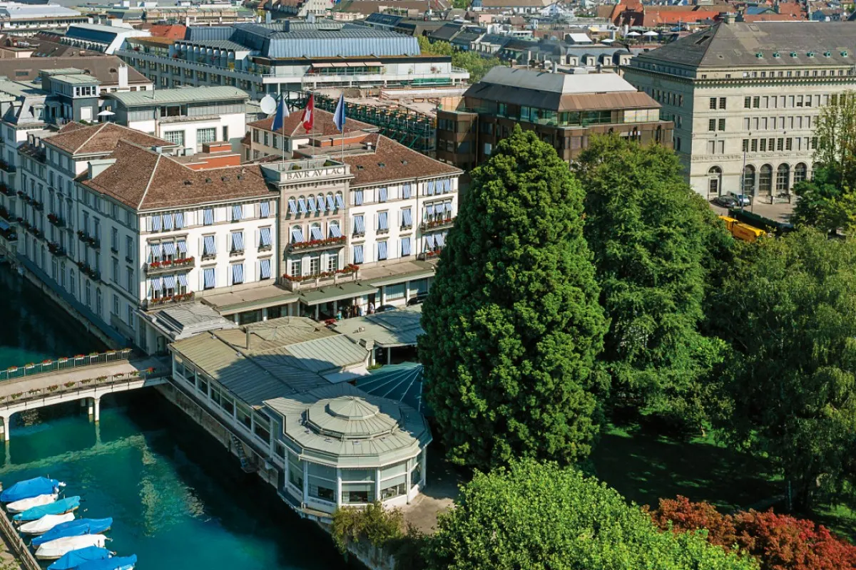 Aerial view of Baur au Lac hotel's riverside façade with flags, striped awnings, gardens and boats on the Limmat.