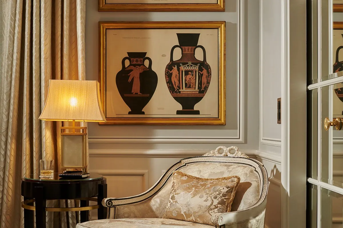 Elegant hotel room corner with framed Greek vase prints, lamp, and upholstered chaise