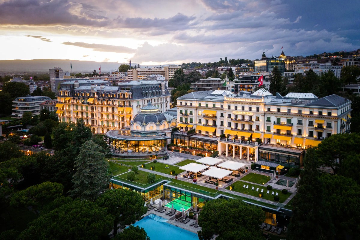 Aerial twilight view of Beau-Rivage Palace hotel with illuminated façade, gardens, terrace and pool, Lausanne cityscape behind