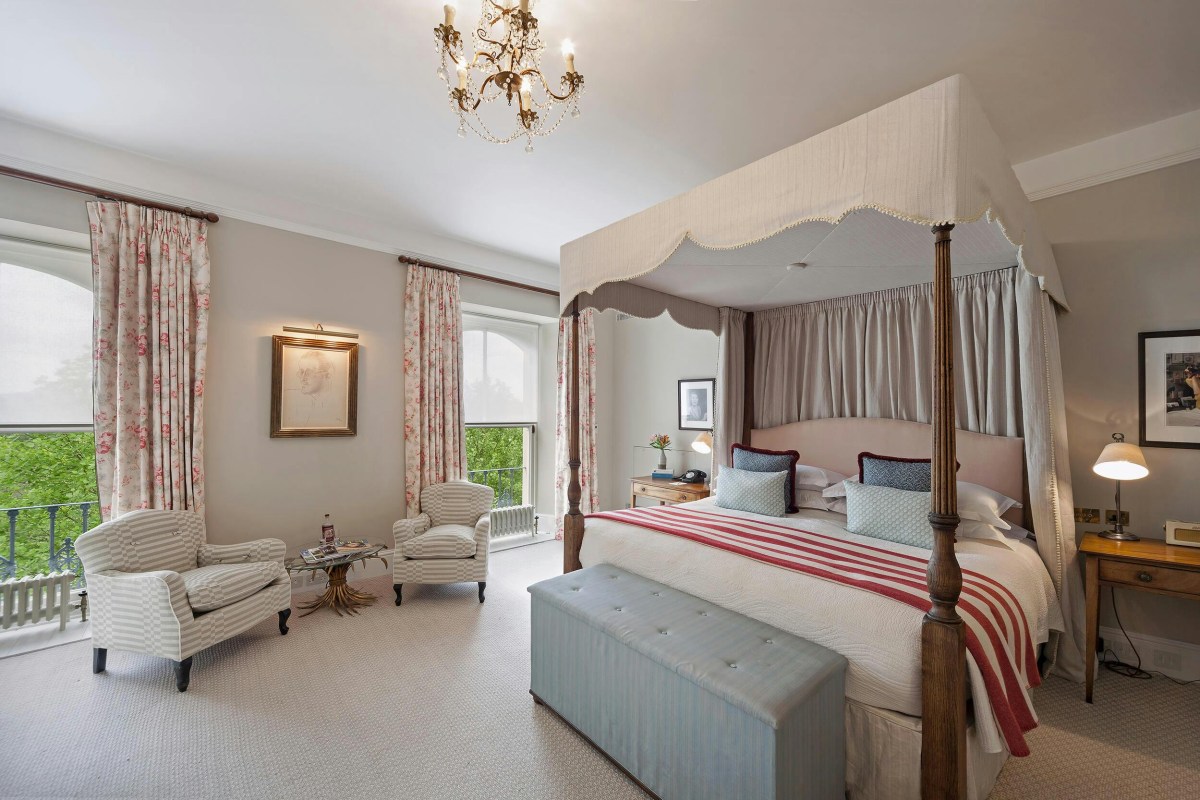 Classic hotel bedroom with four-poster bed, striped armchairs, chandelier and balcony with garden view