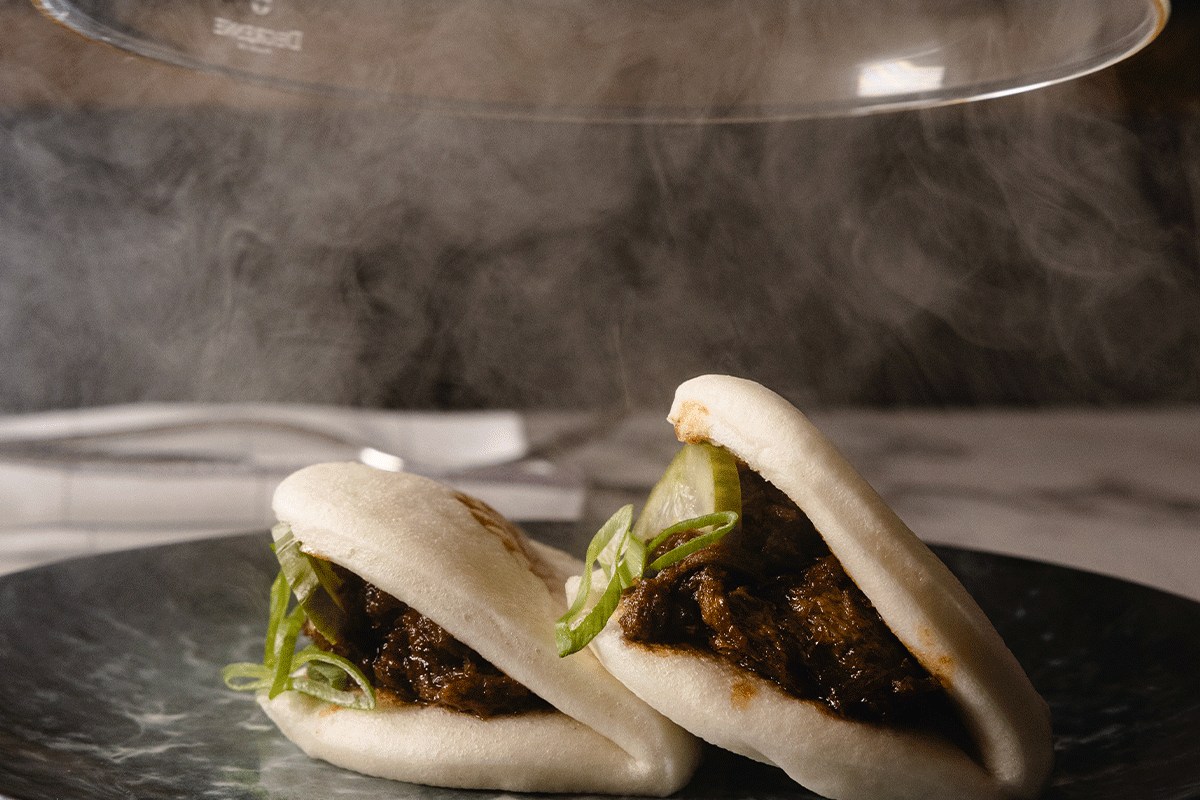 Steamed bao buns filled with braised beef, garnished with scallion and cucumber, served under a smoking dome