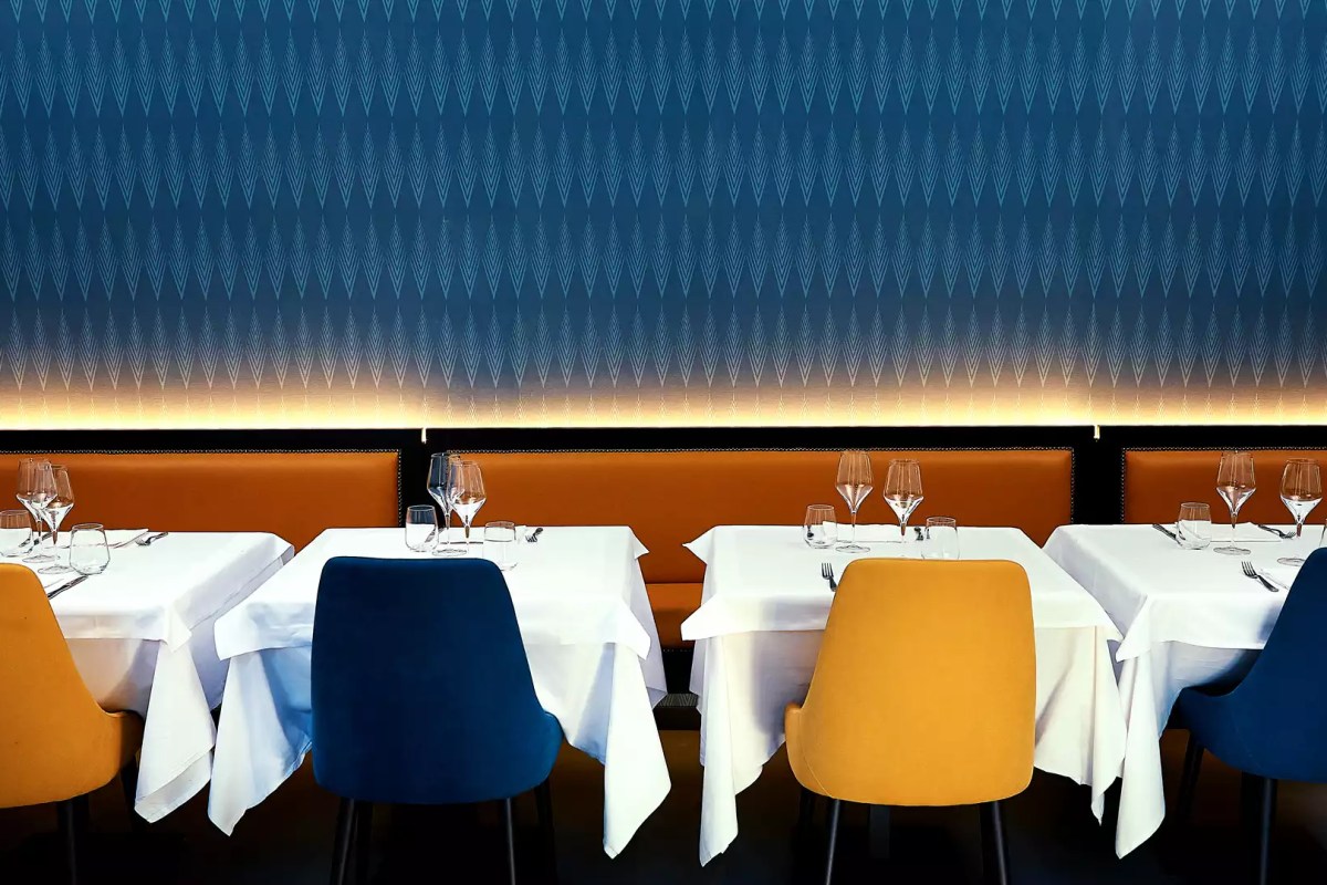 Restaurant dining room with blue patterned wall, orange banquette, blue and yellow chairs, set tables