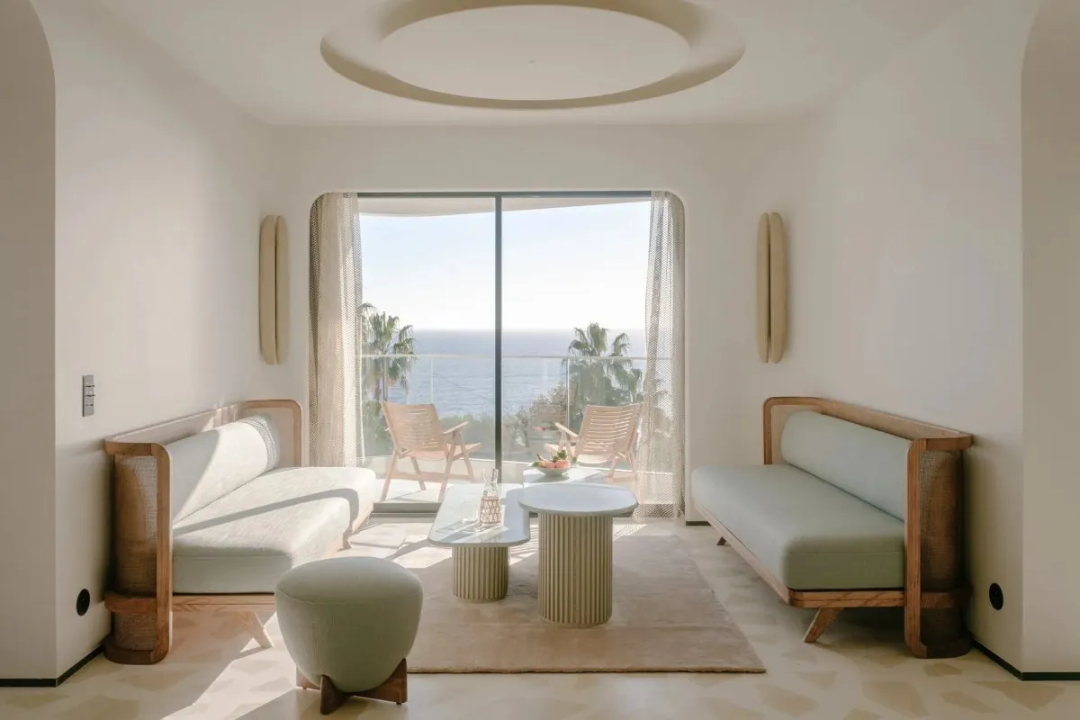 Bright modern suite living area with pastel sofas, round ceiling detail and sea view through curtains