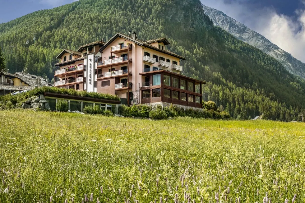 The pink façade of Bellevue Hotel & Spa with its balconies, seen across a summer meadow of wild flowers below wooded mountains in Cogne