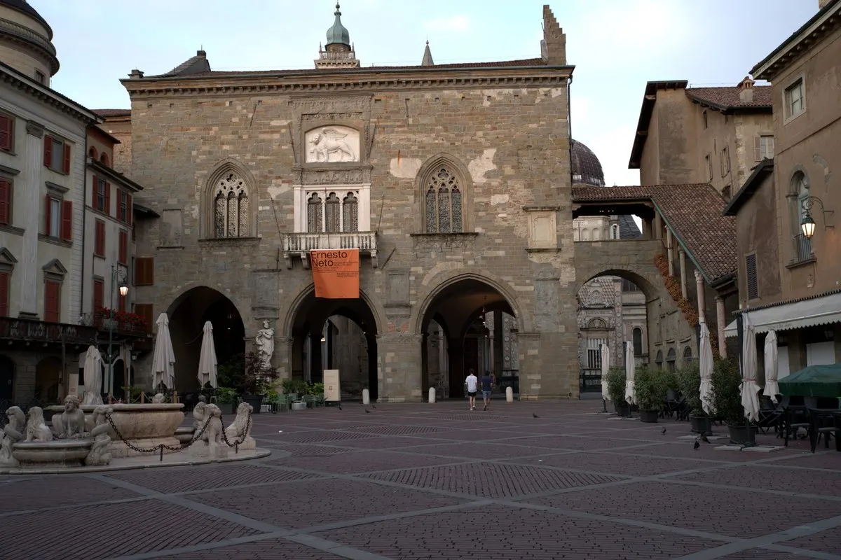 Piazza Vecchia in Bergamo's Città Alta at dusk, with the arcaded Palazzo della Ragione and its Gothic windows, the dome of Santa Maria Maggiore behind and a fountain in the foreground