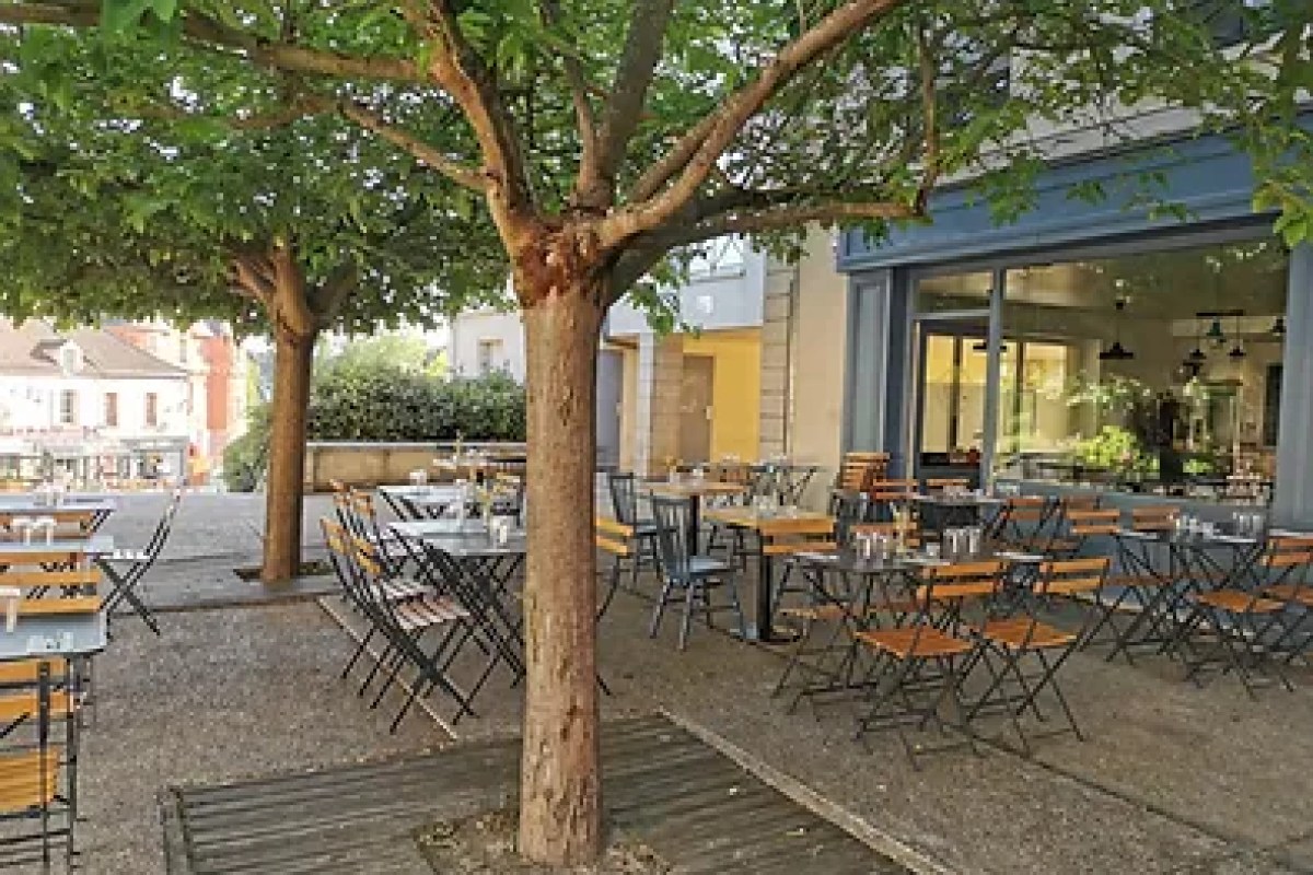 Outdoor terrace of Bird restaurant with wooden tables, chairs, and shade trees beside the building's glass facade.