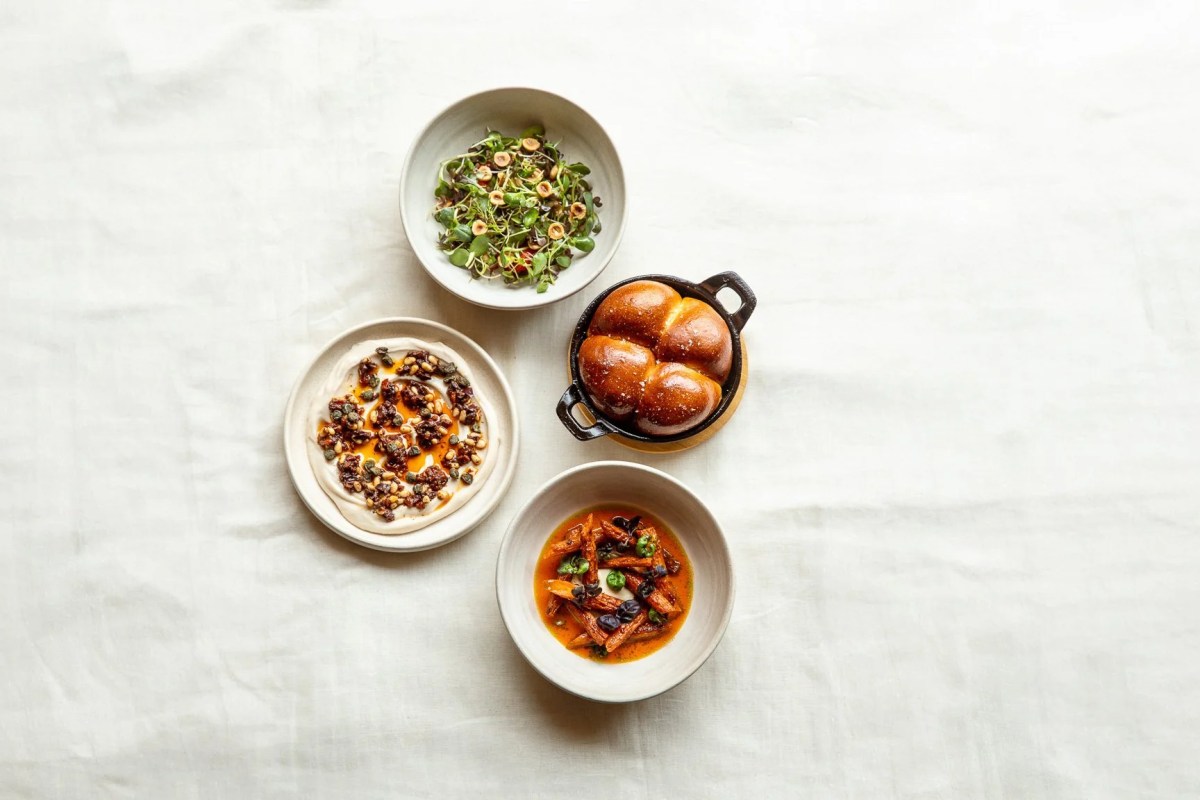 Four small plates on white linen: greens, flatbread with tapenade, bread rolls, and a carrot dish