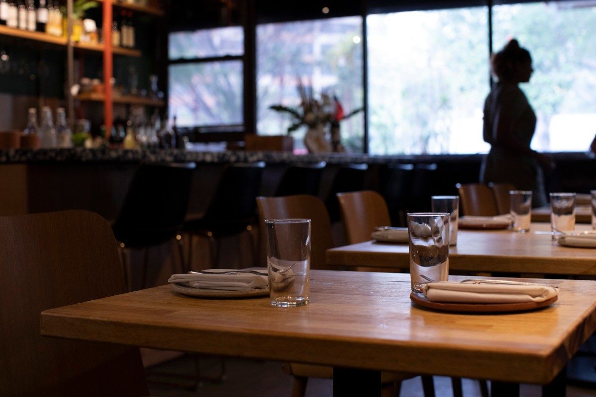 Dining room with wooden tables set with glasses and napkins, bar in background, daylight through windows