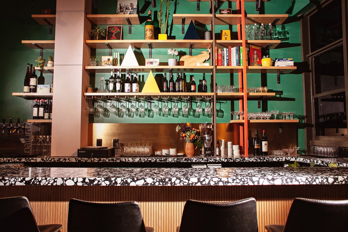 Bar area with terrazzo counter, shelves of wine bottles, glasses hanging, and decorative items against green wall